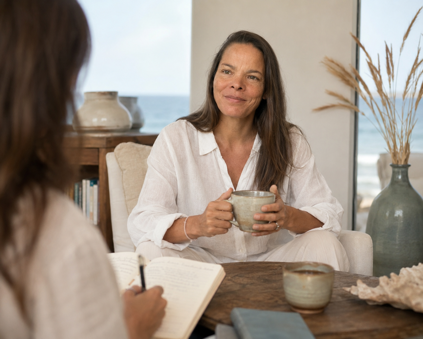 Frédérique, fondatrice de Voie d'Elle, tient une tasse et échange avec une cliente qui prend des notes, dans un salon lumineux face à la mer